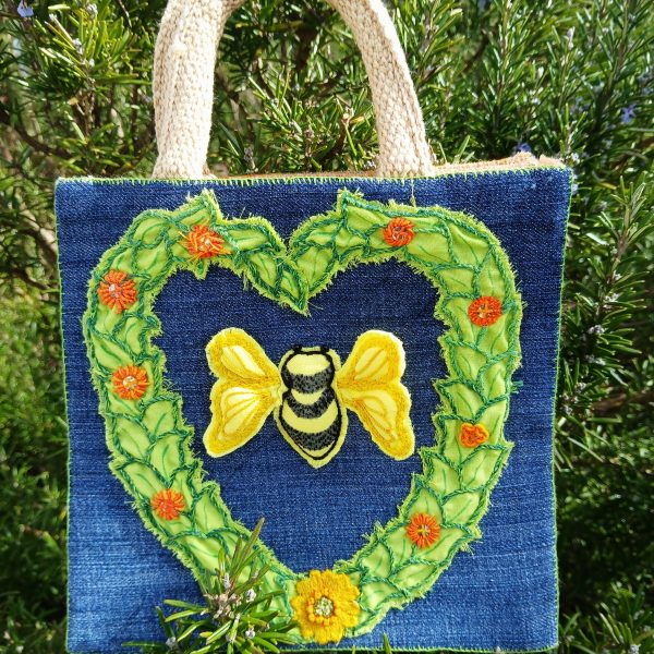 A photograph of a jute shopping bag embroidered with an image of a bumble bee in a heart of leaves and flowers