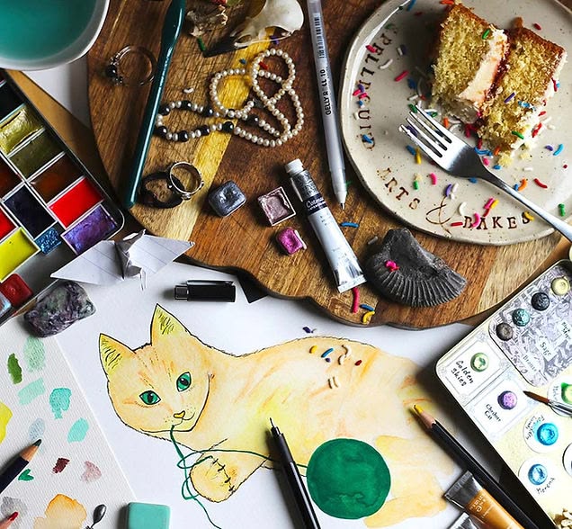 A rich and creative workspace featuring a watercolor painting of a yellow‑orange cat playing with a green ball of yarn. Around the artwork are scattered supplies including paint palettes, brushes, colored pencils, pens, paint tubes, folded paper shapes, jewelry pieces like rings and a pearl necklace, and a small fossil. To one side sits a wooden board holding a plate with crumbs from a partially eaten cake and a fork; the plate is printed with the words “CUPCAKES & BAKES.” The overall scene blends art-making with everyday objects, giving a warm, lived‑in, creative atmosphere.